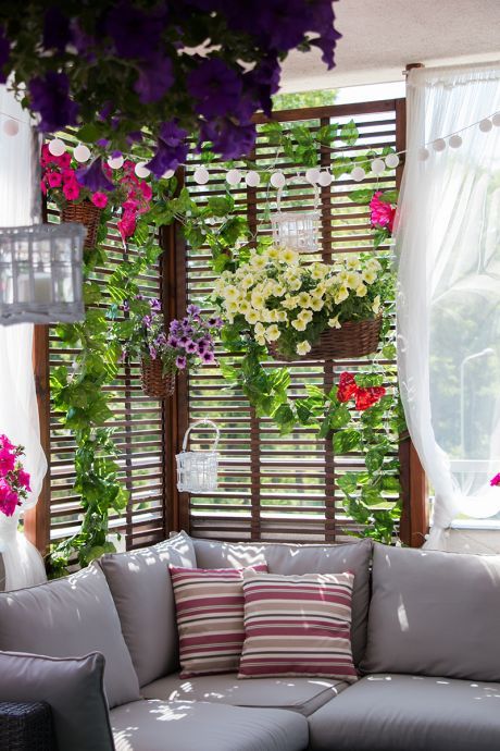 a balcony with a corner sofa and cushions, a trellis with flowers and green plants, some lights and a curtain