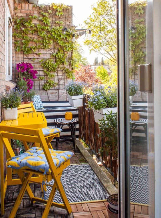 a balcony with potted flowers and green plants, bright yellow furniture, a trellis on the wall and some climbing vines