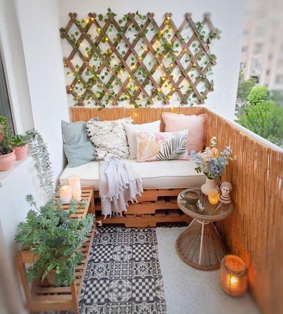 a small cozy balcony with a pallet sofa and cushions, a console table with boards, a side table with flowers and a trellis on the wall with green plants and lights