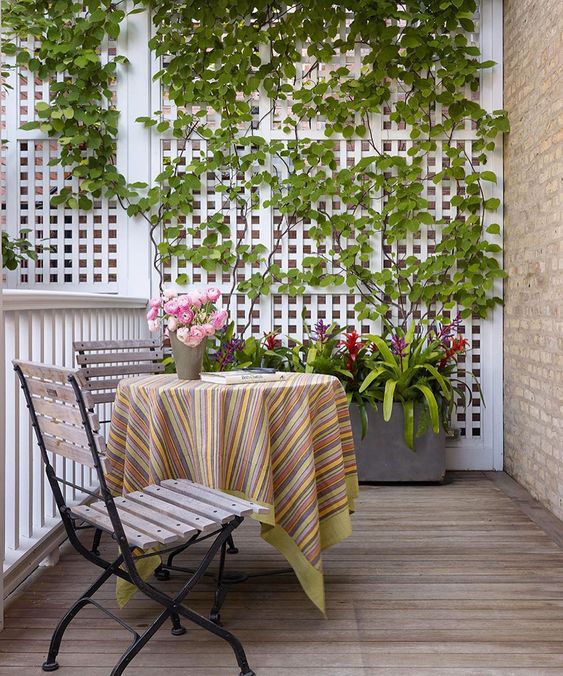 A beautiful and cute balcony with a trellis as a privacy screen with green plants, a planter with flowers, folding chairs and a table as well as some flowers