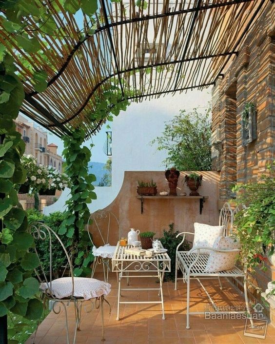 a Provence-inspired balcony with white wrought iron furniture, potted plants and flowers, and a trellis of vines climbing the columns to the roof