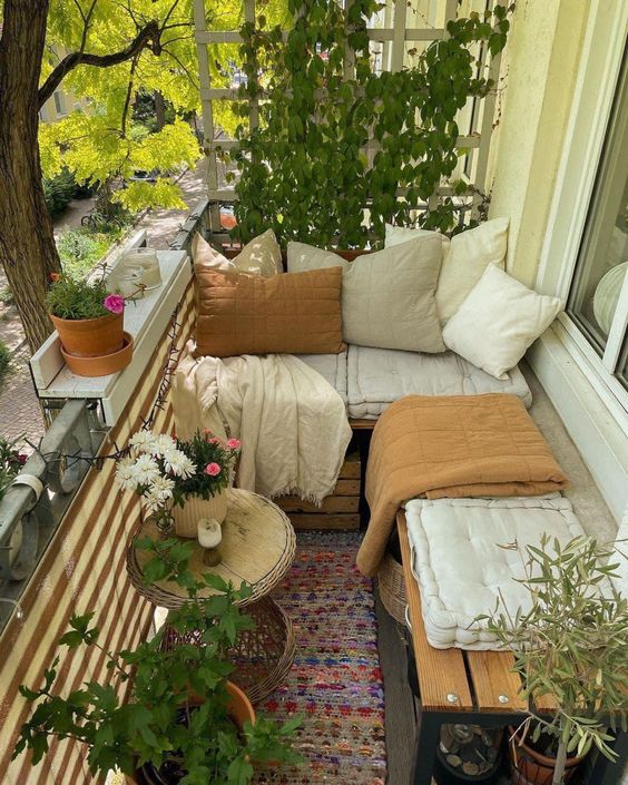 A small and pretty balcony with a love seat and bench, cushions and blankets, side tables, green plants and flowers is fantastic