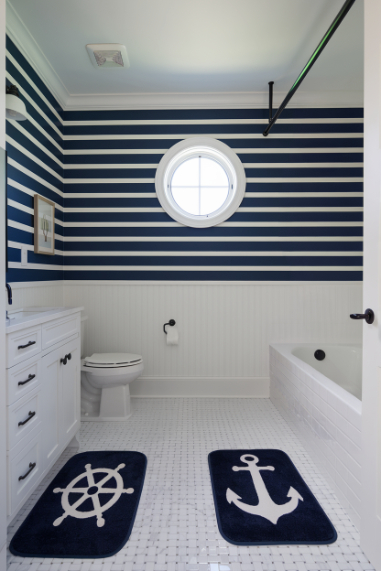 A nautical-themed bathroom with striped walls, a round window and anchor-patterned rugs.