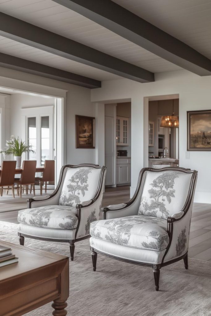 A living room features two patterned armchairs on a neutral rug overlooking a dining area in the background. The room is decorated with framed artwork and a potted plant.