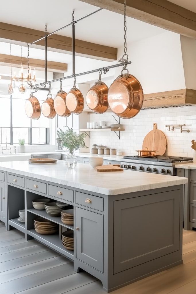 A modern kitchen with a gray island, a marble countertop, copper pots hanging above and a stove with a large cutting board. There are dishes and a vase with green plants on shelves.