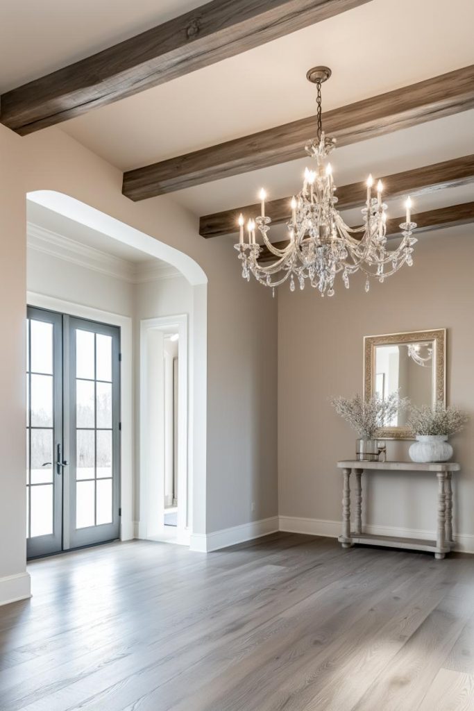 A spacious, light-filled room with exposed beams, a large chandelier and double glass doors. There is a console table with a mirror, vase and decorative items on the wall.