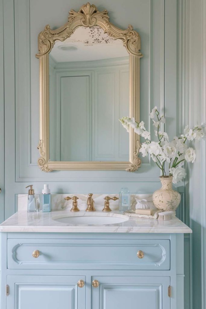 A classic bathroom features a large ornate mirror, a light blue vanity with a marble countertop, gold fixtures, and a vase of white flowers.