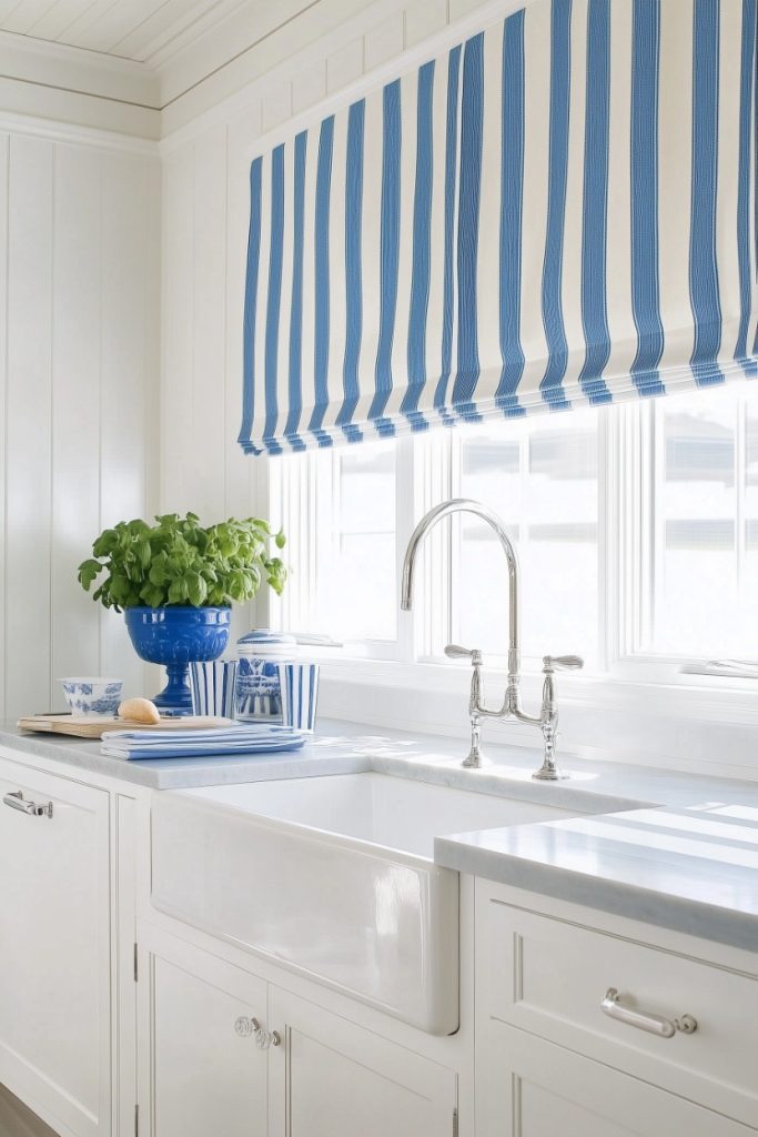 A sink area with a white cabinet and marble countertop, a striped blue and white window blind and a blue vase with green leaves.