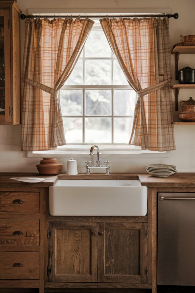 A rustic kitchen features a farmhouse sink, wooden cabinets, checkered curtains, and neatly stacked plates.