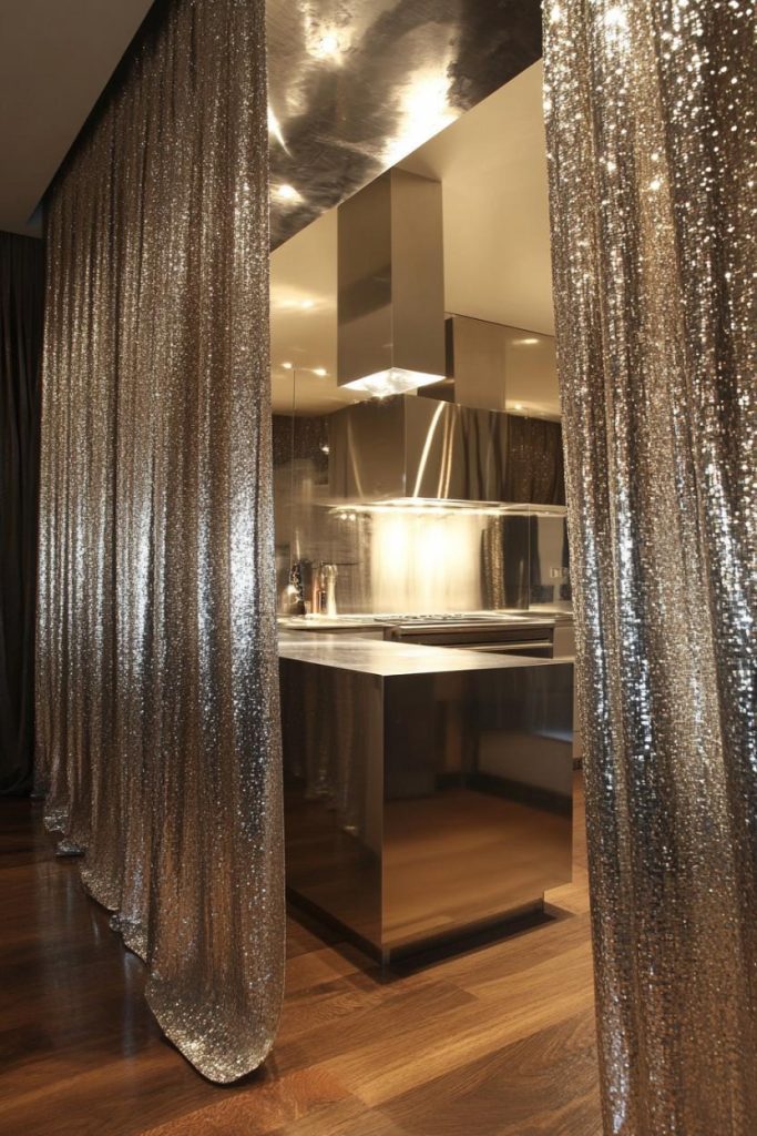 Modern kitchen with metallic surfaces and floor-to-ceiling shimmering curtains. Hardwood floors and sleek stainless steel appliances are visible.