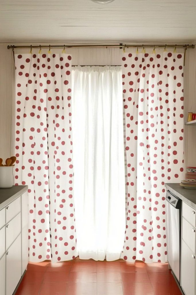 White curtains with red polka dots hang in a kitchen with white cabinets and red tile floors. Natural light penetrates through the transparent inner curtain.