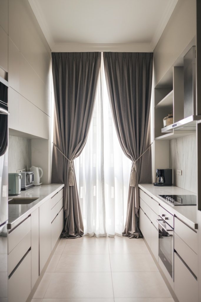 A modern kitchen with white cabinets, built-in appliances and tiled floors. Large windows with gray curtains provide lots of light.