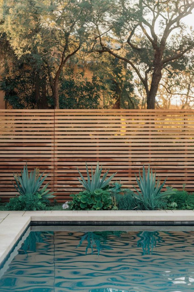 In the foreground a swimming pool with agave plants in front of a wooden picket fence and trees in the background.