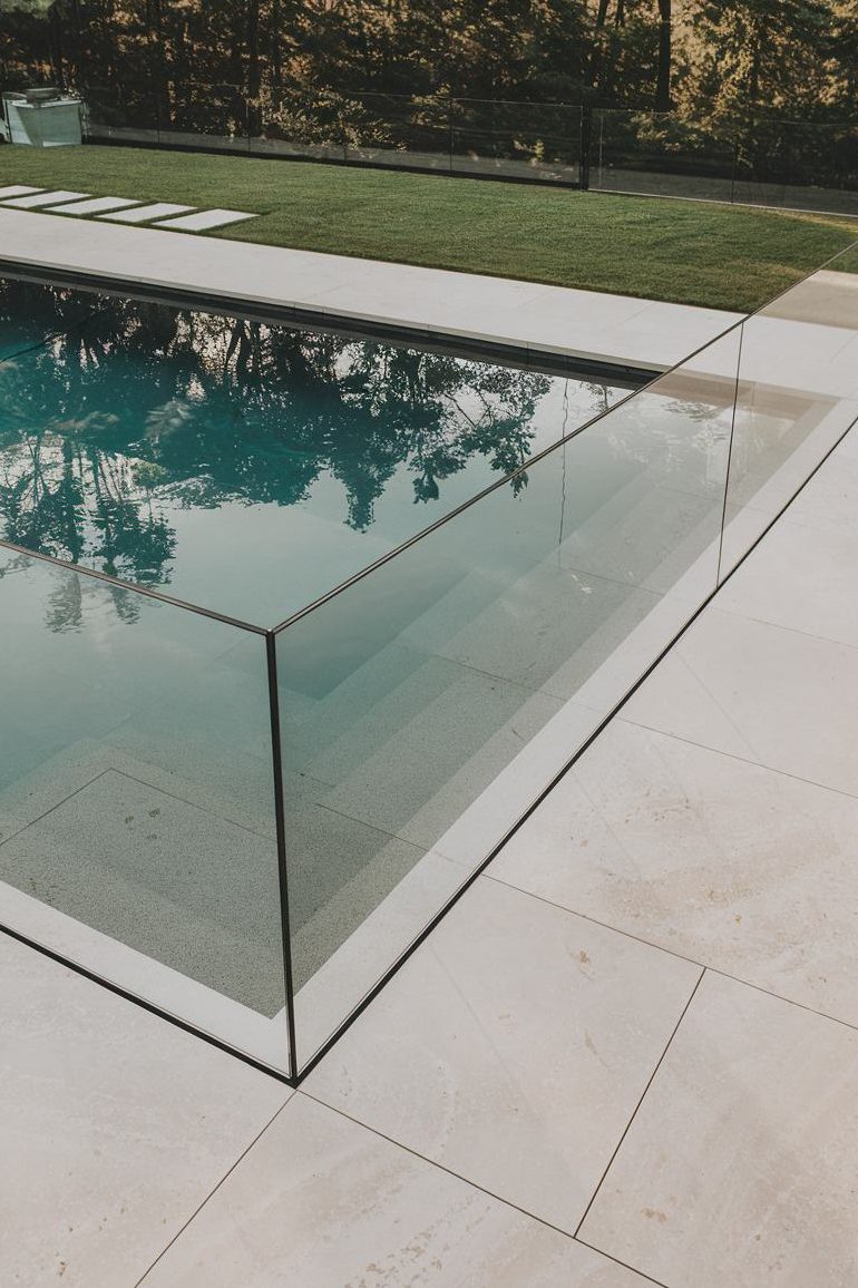 A modern pool with glass walls and clear water is surrounded by light stone tiles. Grass and trees can be seen in the background.