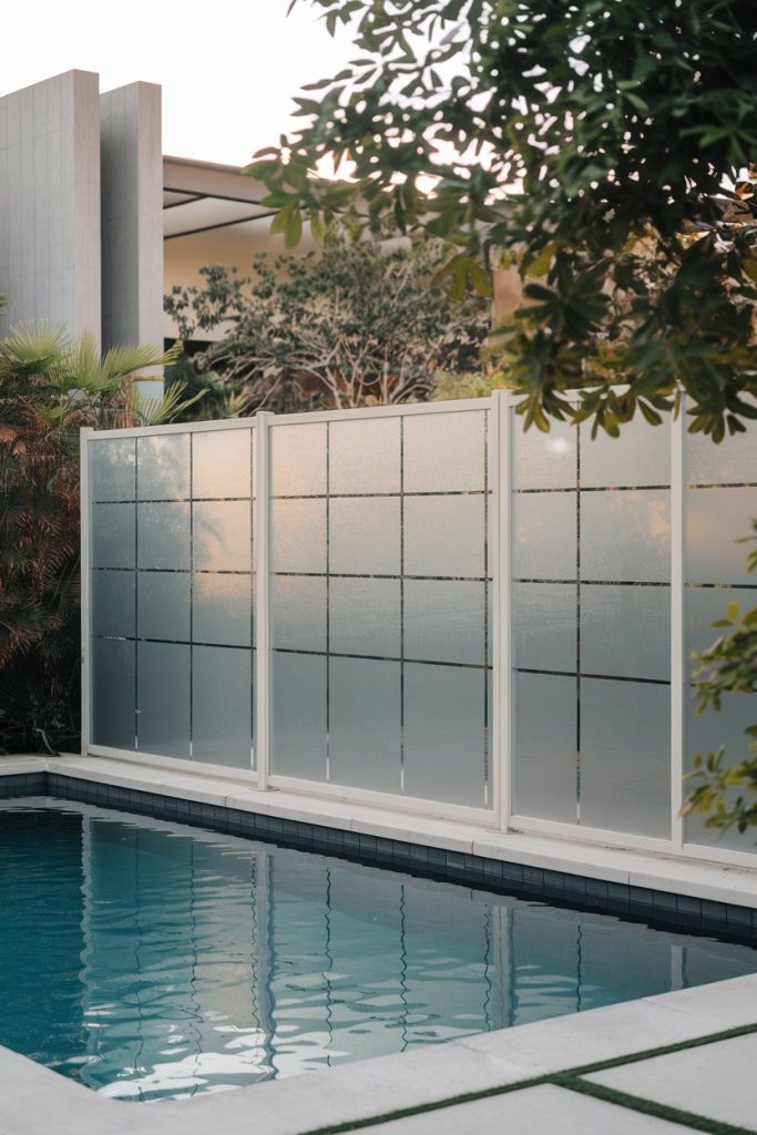 A modern outdoor pool with a glass privacy screen, surrounded by greenery and a modern building in the background.