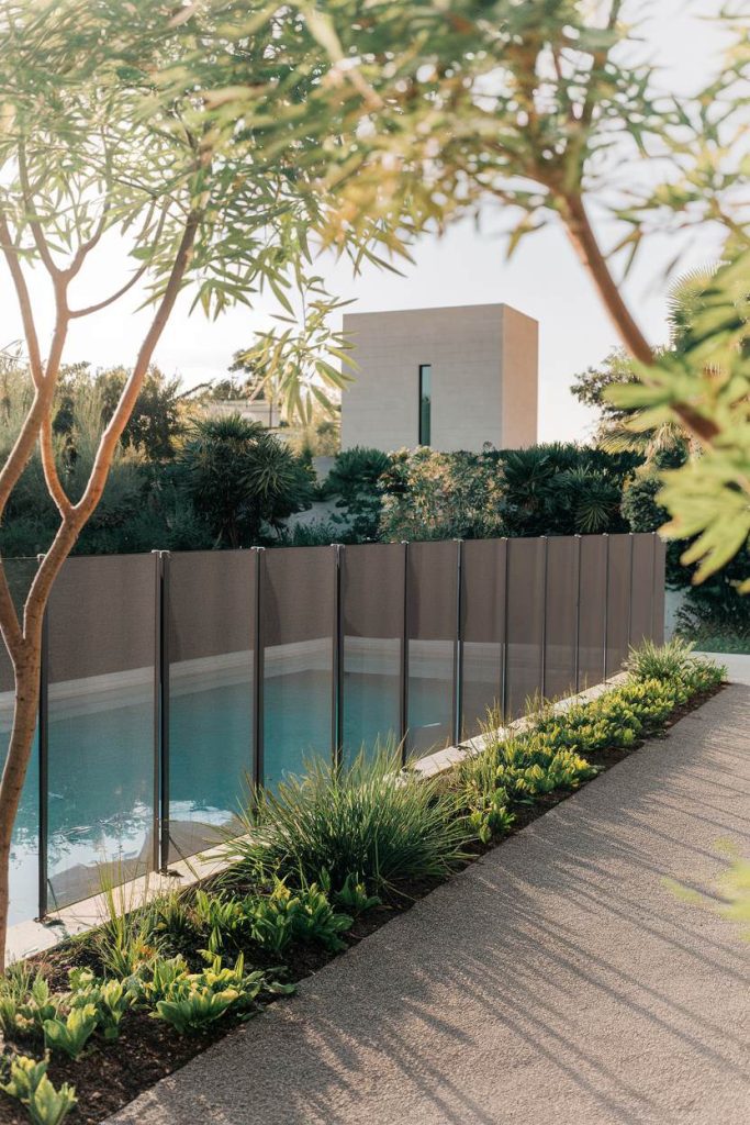 A modern glass fence surrounds a rectangular swimming pool. There is a minimalistic light brown building in the background and the surrounding area is landscaped with lush greenery and trees.