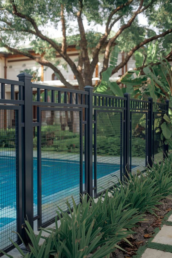 Black metal fence surrounds a garden pool with green plants and a large tree in the background.