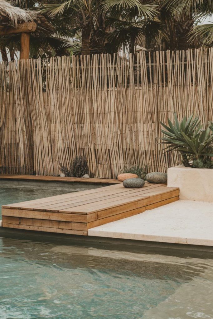 On the terrace by the pool there are two round cushions and a small plant, in the background a bamboo fence and palm trees.