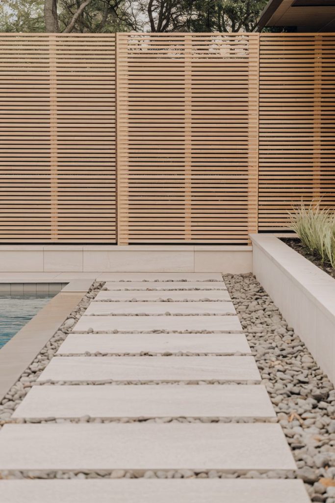 Modern garden path with white stepping stones next to a pool leading to a wooden picket fence and lined with decorative gravel and plants.