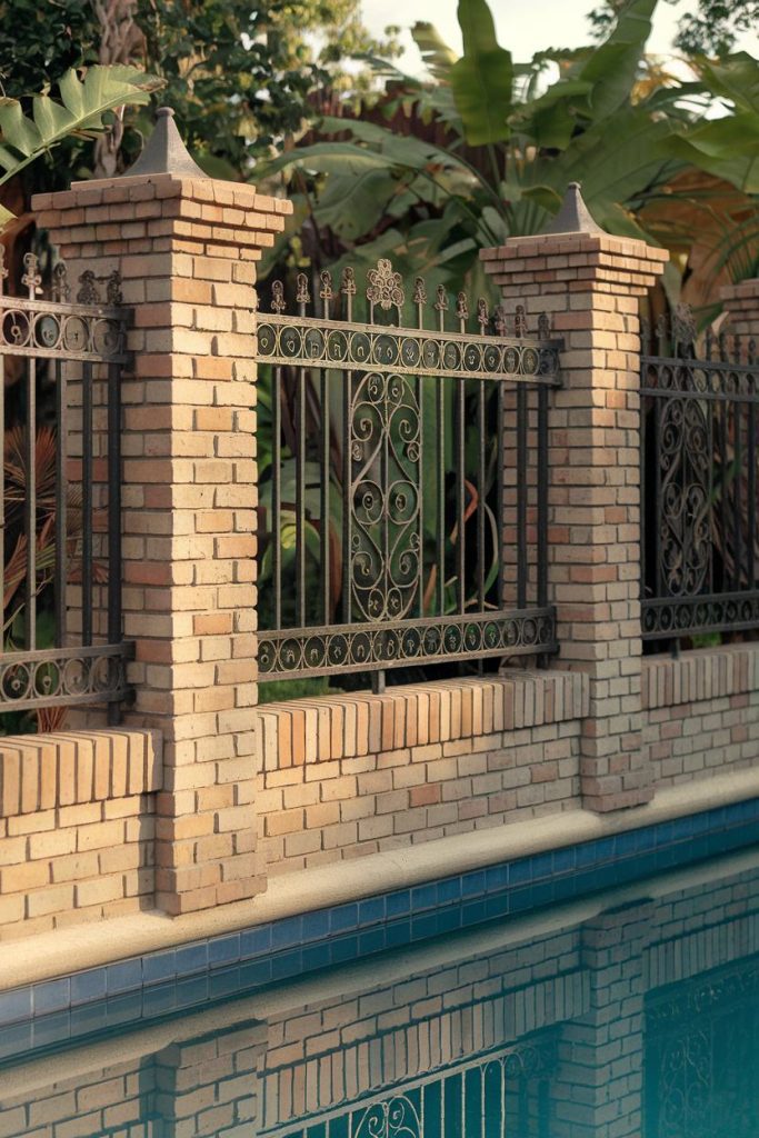 An ornate wrought iron and brick fence surrounds a swimming pool, with lush green foliage visible in the background.