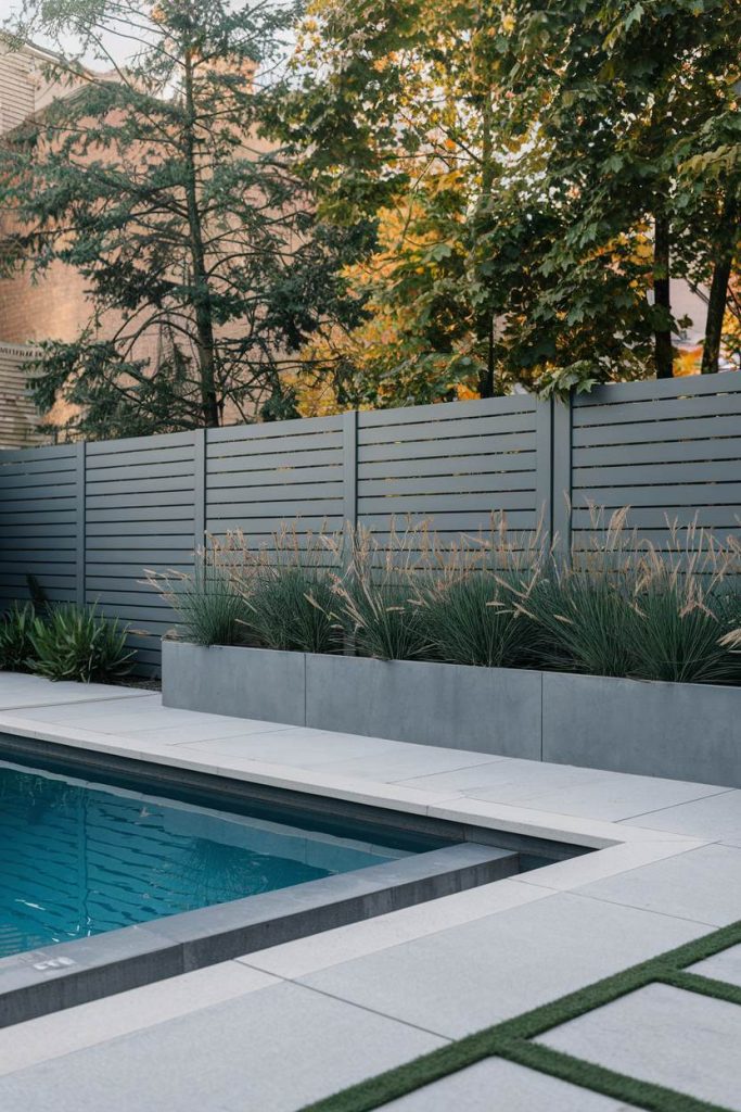 Modern backyard pool with gray fence surrounded by planters with green plants and trees in the background.