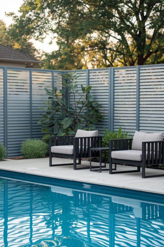 Two black chairs with gray cushions and a small black table next to a clear pool, against a backdrop of gray picket fence and lush greenery.