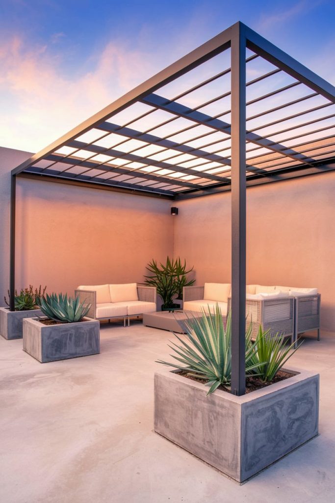 Modern patio with metal pergola, white sofas and large concrete planters with green plants in front of a wall in warm tones under a blue and pink sky.