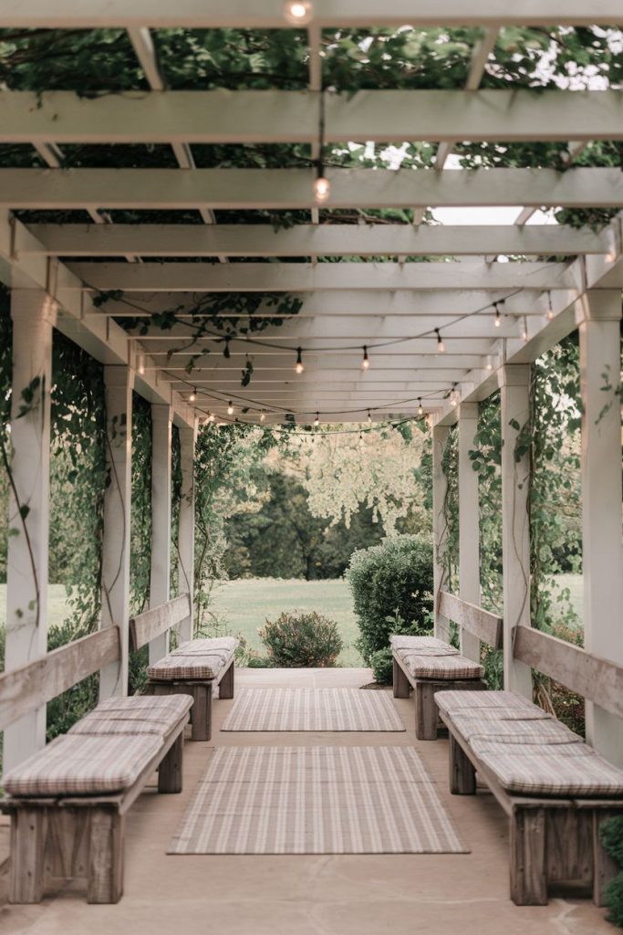 A pergola with fairy lights and vines and benches with cushions on either side. Carpets line the path and greenery can be seen in the background.