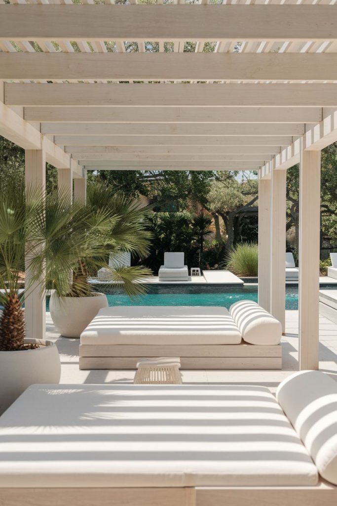 Outdoor lounge area with two cream daybeds under a wooden pergola, overlooking a pool and surrounded by potted plants and greenery.