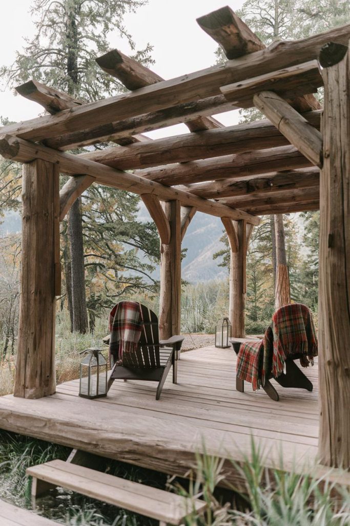 A wooden pergola with two chairs covered in checkered blankets overlooks a wooded landscape. Lanterns are placed next to the chairs.