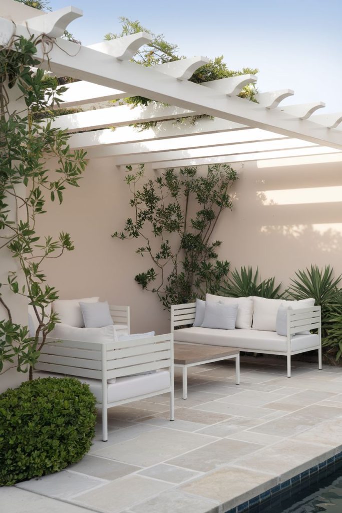 Outdoor terrace with white pergola, two white upholstered sofas, green plants and tiled floor by the pool.