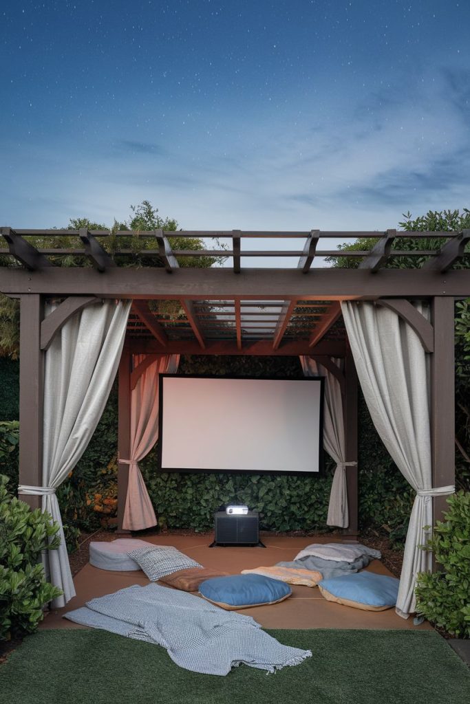 Outdoor film setup under a pergola with a projector screen, curtains, cushions and a small table. The scene is surrounded by greenery under a starry sky.