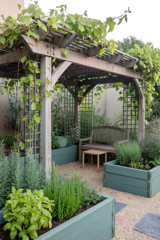 A wooden pergola covered in vines stands above a bench, surrounded by raised beds full of various plants. Gravel paths lead through the garden area.