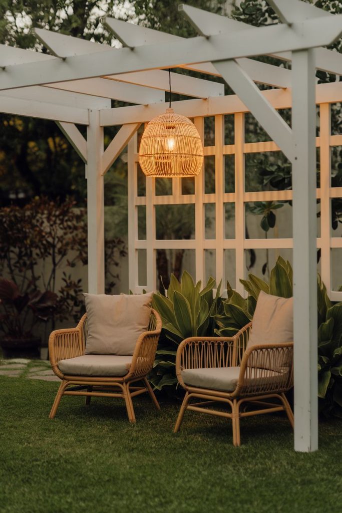 Outdoor seating area with two wicker chairs under a white pergola and an illuminated woven pendant lamp, surrounded by greenery.