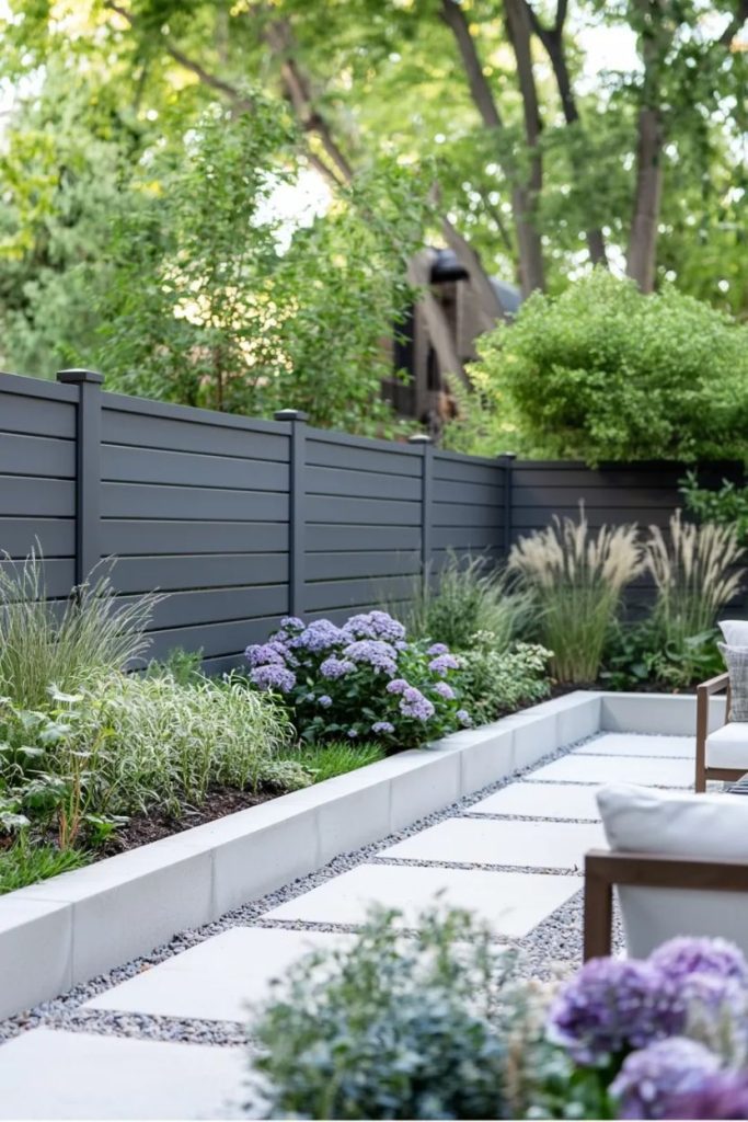 Modern garden with gray fence, lush greenery, purple and white flowers and a clean stone path. Chairs can be seen in the foreground.