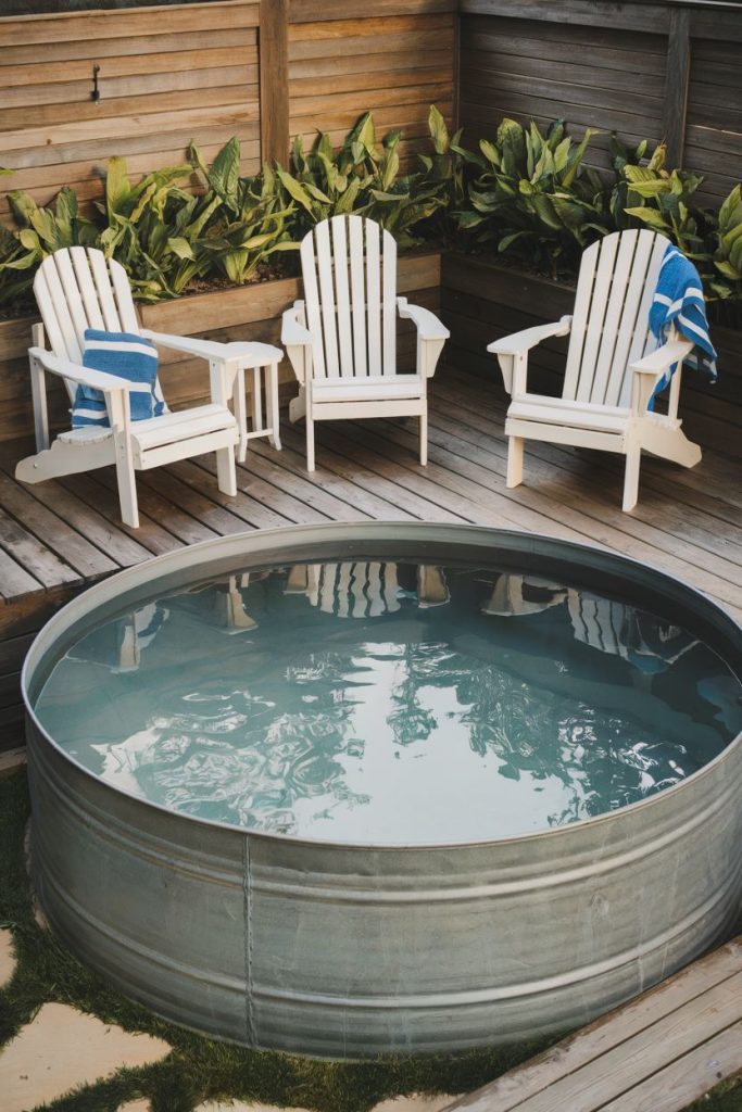 Round metal storage tank next to a wooden patio with two white Adirondack chairs and green plants.