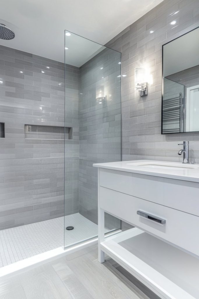 Modern bathroom with walk-in glass shower, wall lamp, rectangular mirror and white vanity unit with sink. Gray tiled walls and light wooden floors complete the design.