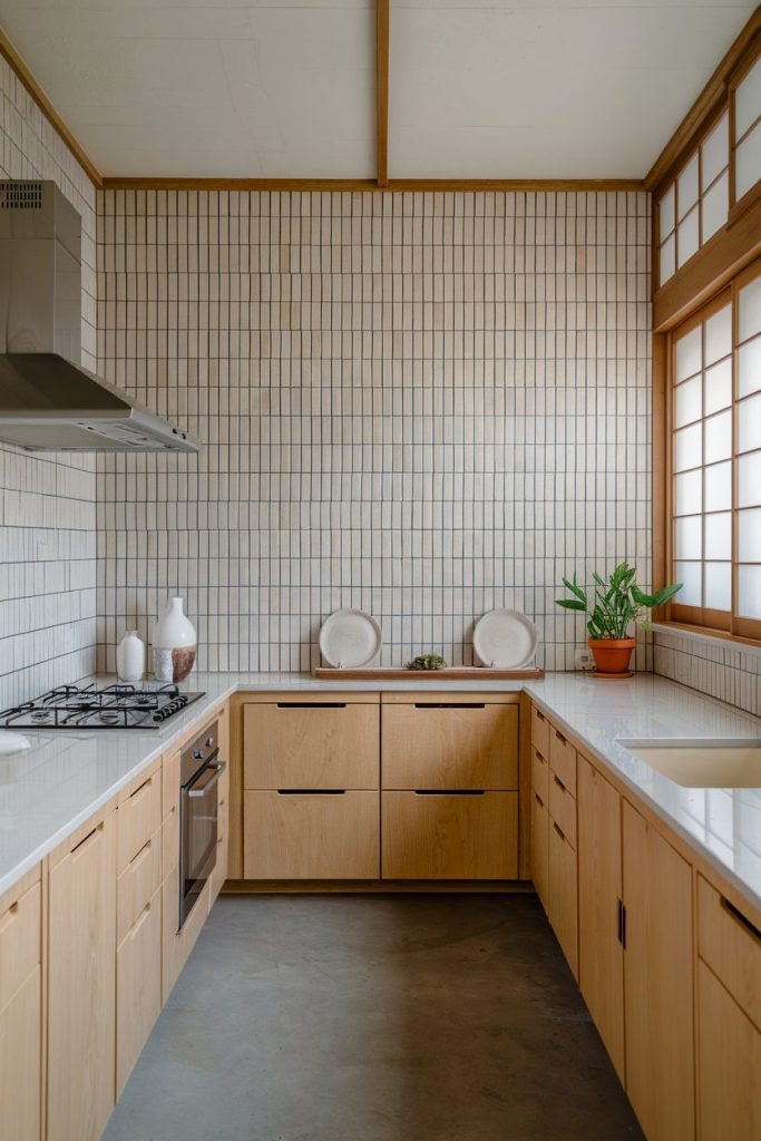 Minimalist kitchen with light wooden cabinets, a gas stove and white countertops. Tiled walls, a potted plant and a small window with wooden paneling complete the furnishings.