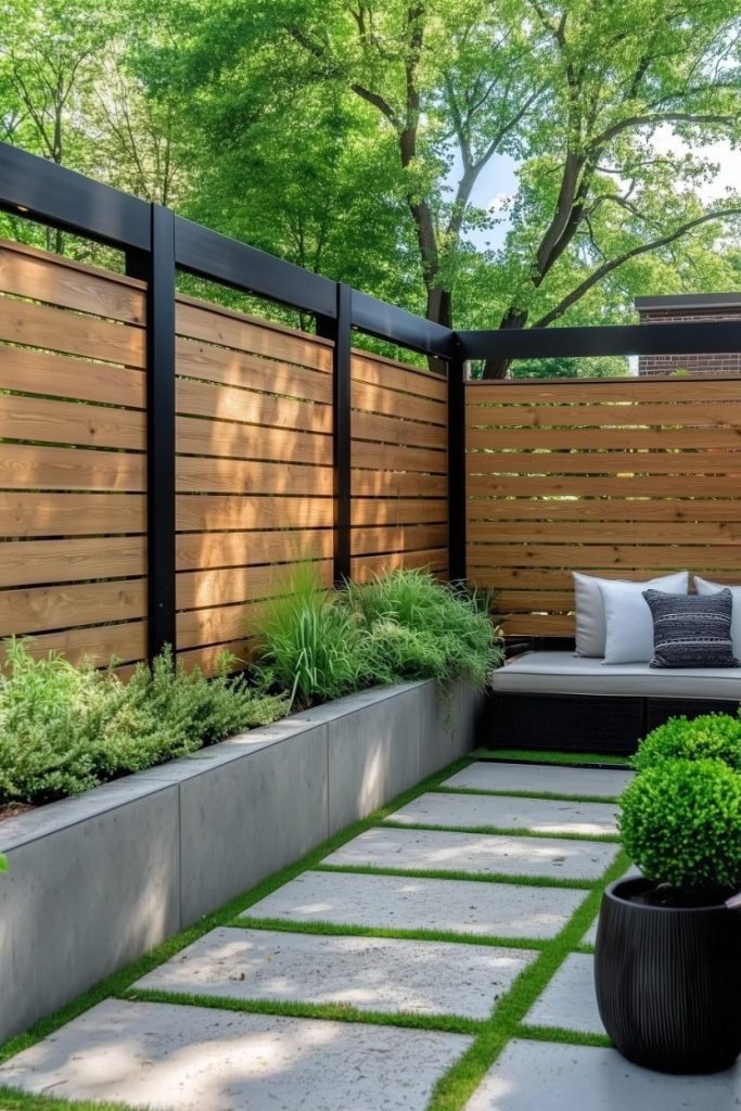 Modern patio with wooden privacy fence, concrete paving stones and planters. Greenery lines the perimeter and there is a cushioned seating area in the corner.