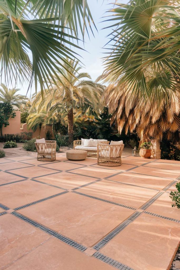 A sun-drenched terrace with wicker chairs and a table surrounded by palm trees and greenery.