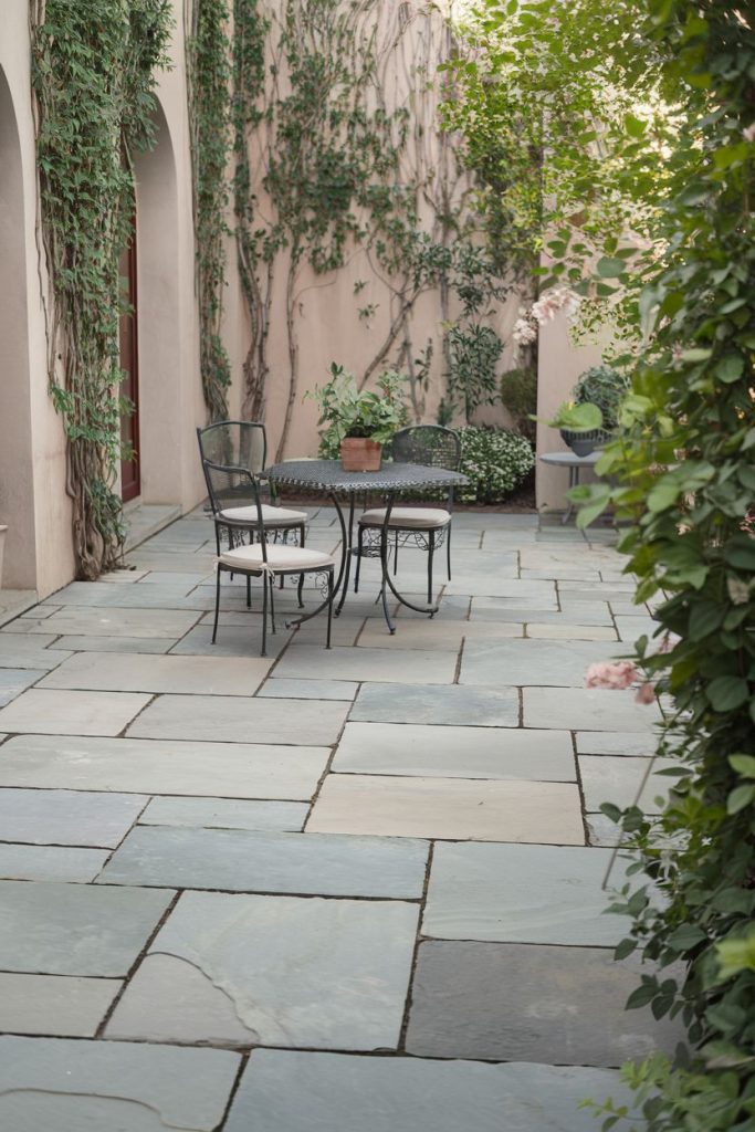 A stone-tiled patio features a metal table and two chairs surrounded by planted walls and greenery.