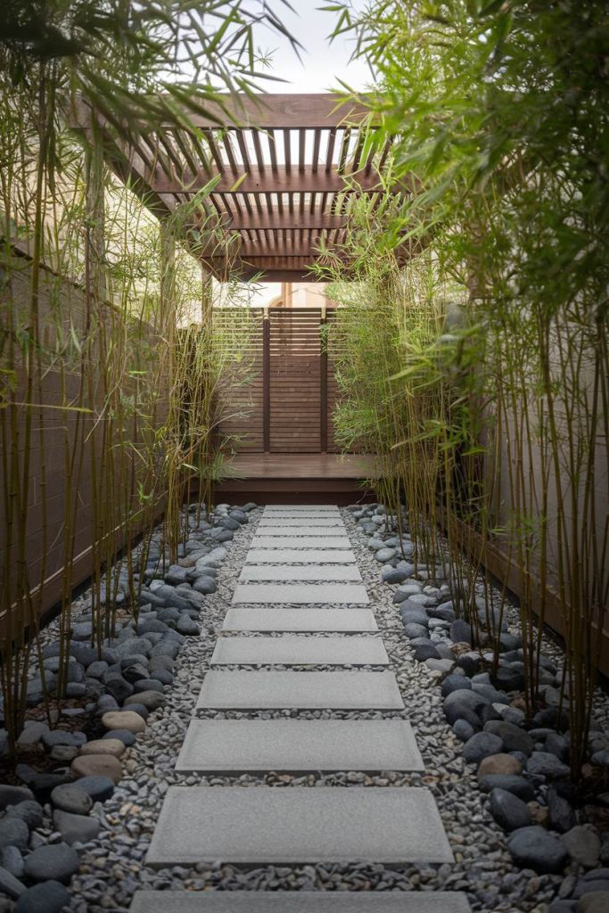 A stone path lined with bamboo plants leads to a wooden gate under a pergola.