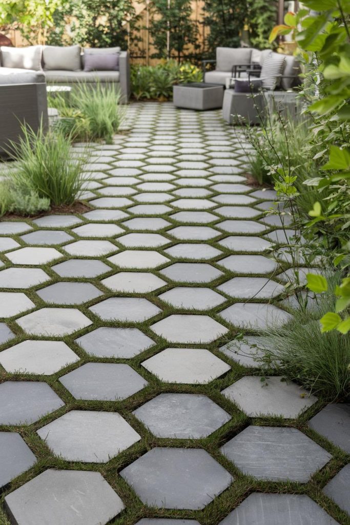 Hexagonal stone tiles with grass in between form a path leading to outdoor seating with gray cushions and surrounding greenery.