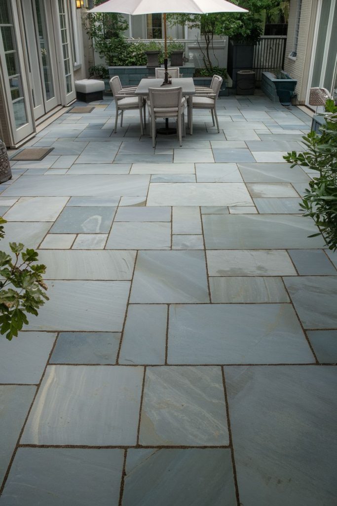 A gray stone tiled terrace furnished with a dining table and chairs under a large white parasol surrounded by potted plants.