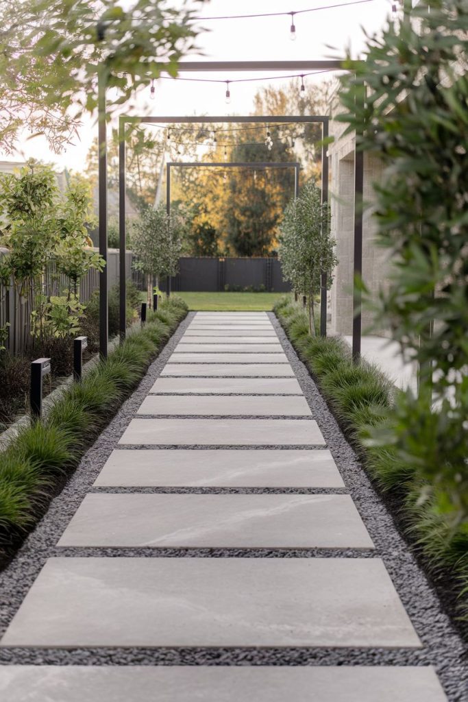 A modern garden path with large rectangular stone slabs and surrounding greenery. Metal arches stretch above it and fairy lights are visible. A fence and trees can be seen in the background.