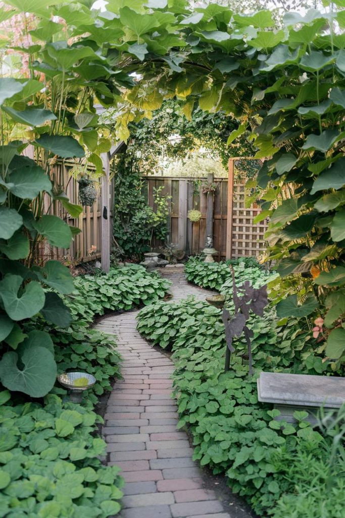 A brick path winds through a lush garden with dense greenery and leads to a wooden fence. A metal sculpture and a bench can be seen on the right.