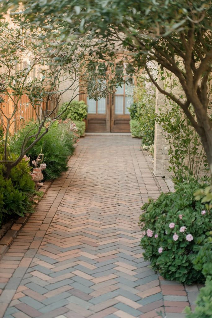 A brick herringbone path lined with green plants and pink flowers leads to a set of wooden doors. Trees frame the entrance.