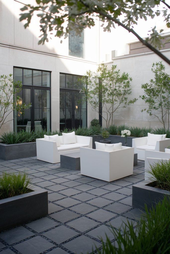 Modern outdoor patio with white square chairs and tables surrounded by rectangular planters with green plants. Large windows and bright building walls in the background.