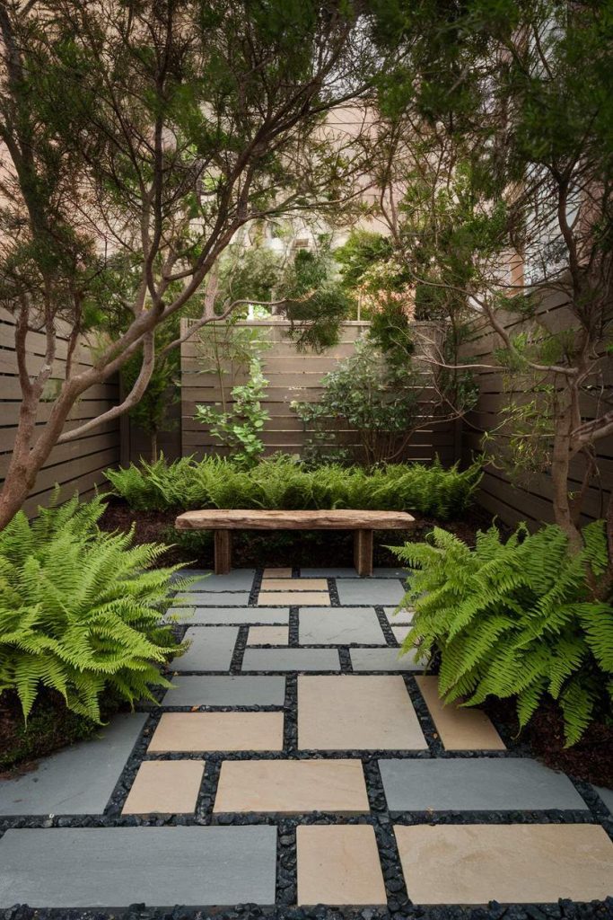 A tranquil garden features a stone path leading to a wooden bench, surrounded by ferns and trees and surrounded by a wooden fence.