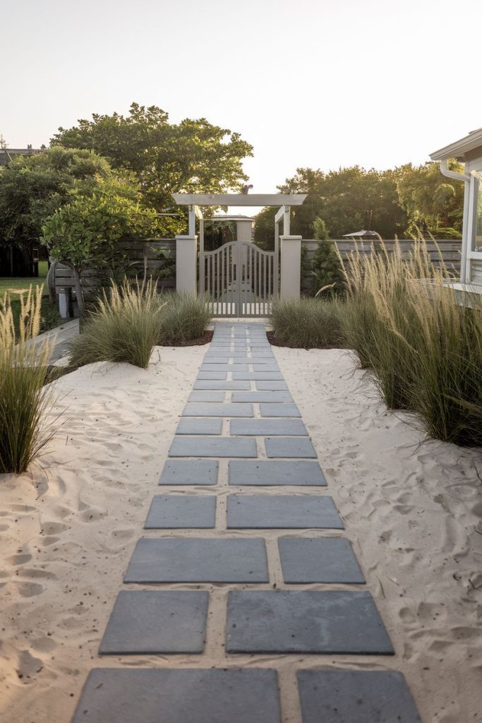 A path made of square stones leads through a sand-filled garden with tall grasses and ends at a white gate under a pergola surrounded by greenery.